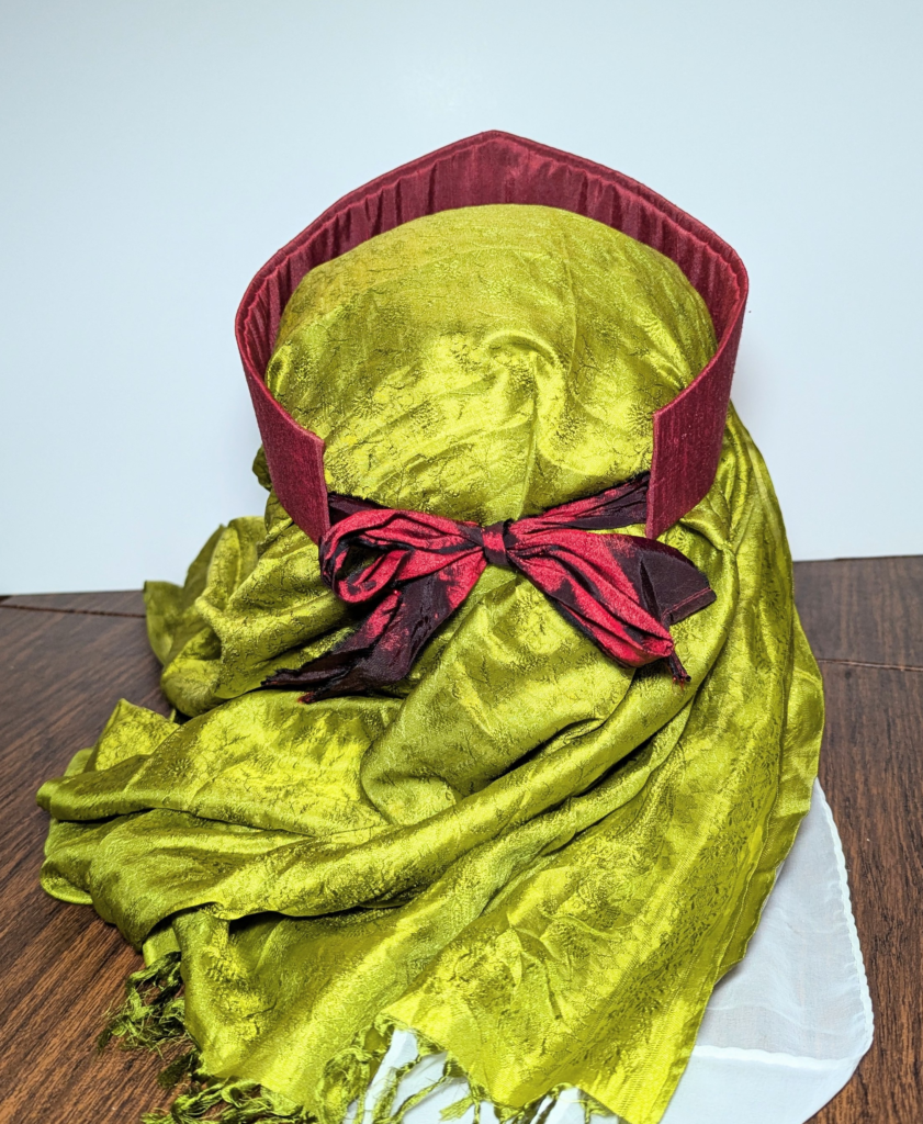 Back view of a canvas mannequin head wearing a red hat over a green scarf and a white veil